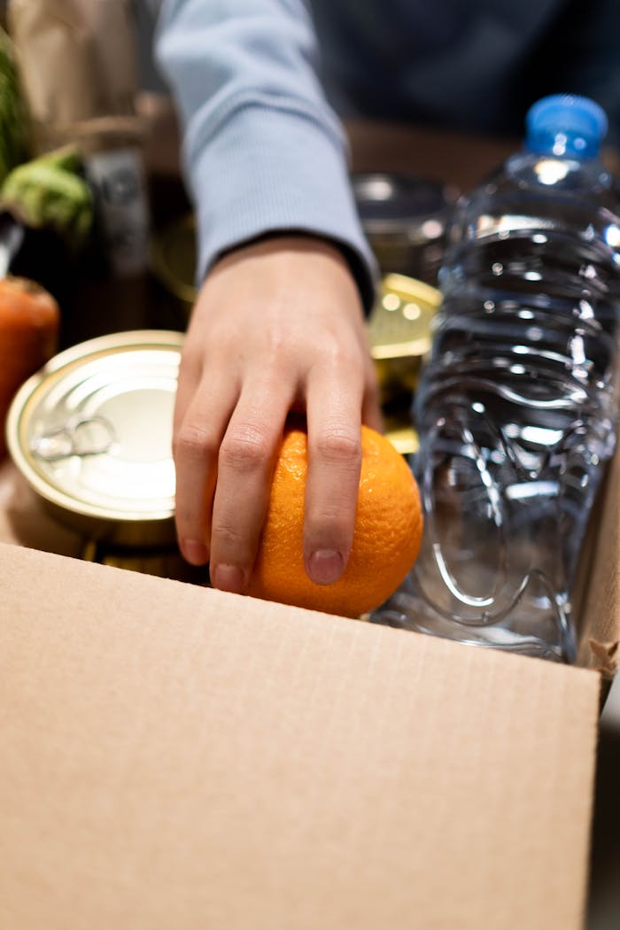 A hand packing an orange, canned food, and a water bottle into a box, symbolizing aid.