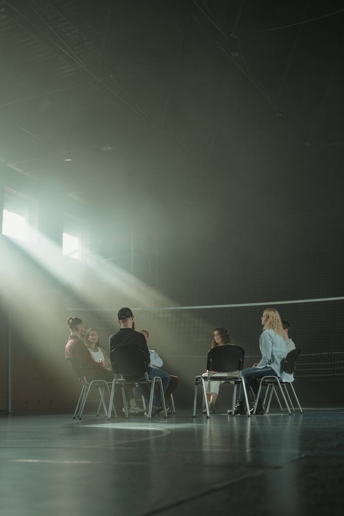 A group of adults in a therapy session, sitting in a dimly lit room with sun rays streaming in.